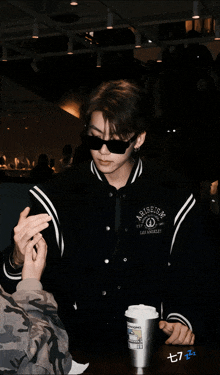 a man wearing sunglasses and a jacket that says arise on it sits at a table with a cup of coffee