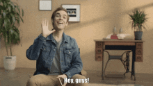 a man in a denim jacket says hey guys in front of a desk
