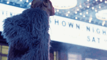 a woman in a blue fur coat stands in front of a sign that says shown night sat