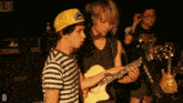 a man wearing a yellow trucker hat holds a guitar next to another man
