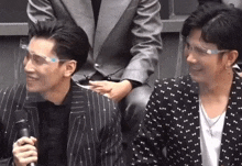 two men in suits and glasses are sitting next to each other . one of the men is holding a microphone .