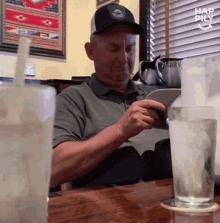 a man in a hat sits at a table reading a book and drinking a glass of water
