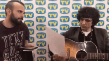 two men are playing guitars in front of a wall with tv logos