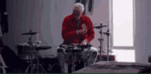 a man in a red jacket is playing a drum set in a room .