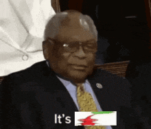a man in a suit and tie is sitting in front of a sign that says `` it 's '' and a flag .