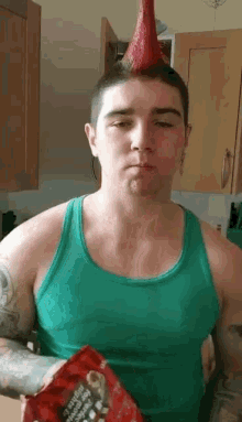 a man wearing a green tank top and a red party hat holds a bag of popcorn