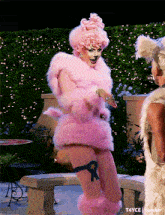 a drag queen is wearing a pink fur coat with a blue ribbon on her leg