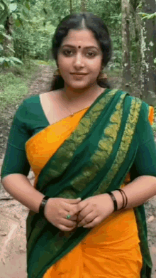 a woman in a green and yellow saree is standing on a path in the woods .