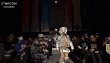 a woman in a white mask stands in front of a crowd with a stardom logo in the background