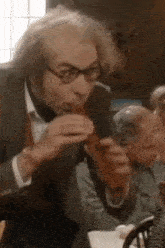 a man in a suit and tie is eating a hot dog while sitting at a table .