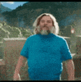a man with long hair and a beard is wearing a blue shirt and standing in front of a mountain .