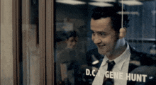 a man in a suit and tie is smiling in front of a window that says d.c. gene hunt on it