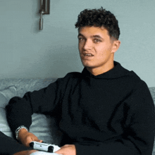 a young man in a black sweatshirt is sitting on a couch holding a video game controller