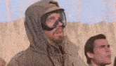 a man wearing a hooded jacket and goggles is standing next to another man in the desert .
