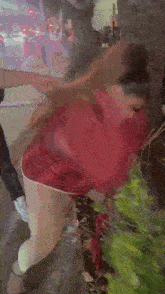 a woman in a red shirt and white pants is standing in front of a bush