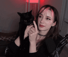 a woman with pink hair and a choker holds a black cat
