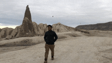 a man in a black jacket stands in the desert in front of a large rock formation