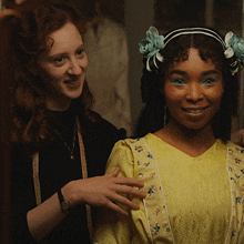 a woman wearing a yellow dress with flowers in her hair smiles next to another woman
