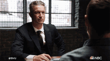 a man in a suit and tie sits at a table talking to another man with a nbc logo in the corner