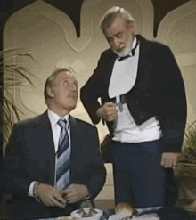 a man in a tuxedo talks to another man in a suit
