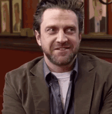 a man with a beard is wearing a brown jacket and a white shirt .