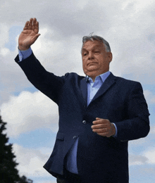 a man in a blue suit is waving his hand in front of a cloudy sky