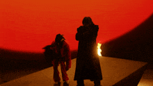 a man in a red jacket stands next to a man in a black coat in front of a fire