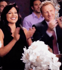 a woman in a black dress applauds while a man in a suit looks on