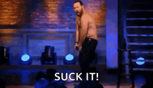 a man without a shirt is standing on a stage with a microphone in his hand and says suck it .