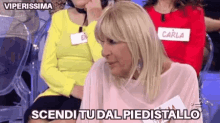 a woman with blonde hair is sitting in a chair and says scendi tu dal piedistallo