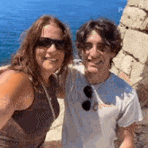 a man and a woman are posing for a picture in front of the ocean . the woman is wearing sunglasses .
