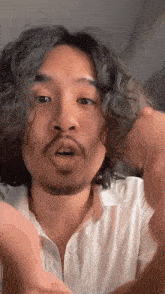 a man with curly hair and a beard is wearing a white shirt and making a funny face .