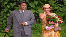 a man in a suit holds a bicycle while a woman in a hat holds flowers