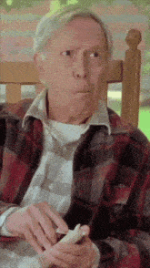 a man in a plaid shirt sits on a porch eating a sandwich