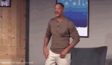 a man in a brown shirt and white shorts is dancing in front of a couch and a picture .