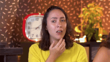 a woman in a yellow shirt is making a face in front of a clock that shows the time as almost 5:00