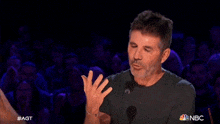 a man in a black shirt stands in front of a microphone and applauds in front of a nbc logo