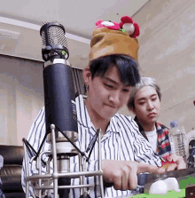 a man wearing a hat is cutting something in front of a microphone while another man looks on .