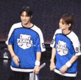 two men wearing blue and white teams jerseys are standing next to each other on a stage .