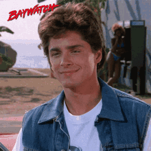 a man wearing a denim vest is smiling in front of a baywatch poster