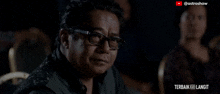 a man with glasses is sitting in a dark room with a caption that says macam ni pun