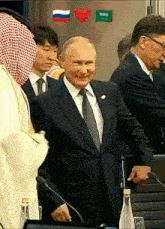 a man in a suit and tie stands in front of a microphone with a heart and saudi flag on the wall behind him