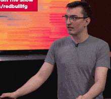 a man wearing glasses and a grey shirt stands in front of a screen that says " join the show "