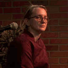 a woman wearing glasses stands in front of a microphone in front of a brick wall