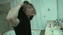 a man in a black shirt adjusts his hair in front of a bathroom mirror with the letter d above him