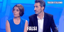 a man in a suit and a woman in a blue dress are sitting next to each other and the man is saying falsi