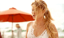 a woman in a white tank top is standing on a beach with an umbrella in the background .