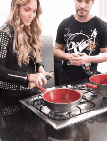 a man and a woman are cooking on a stove and the woman is wearing a black jacket with studs
