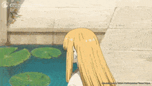 a girl with long blonde hair is standing in front of a pond with lily pads ..