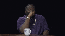 a man in a purple shirt sits at a table with a white mug that says blue